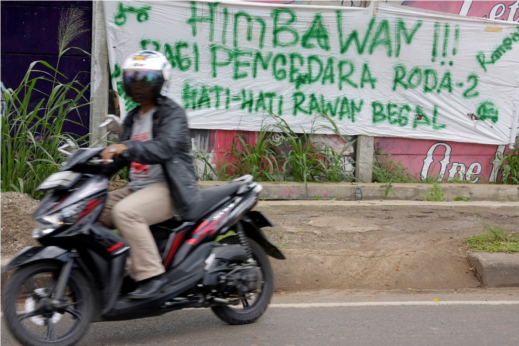 Pengendara motor melintas di dekat spanduk himbauan bagi pen gendara roda dua hati-hati rawan begal dan rampok di Jalan Juanda, Depok. (Foto:MI/Barry)
