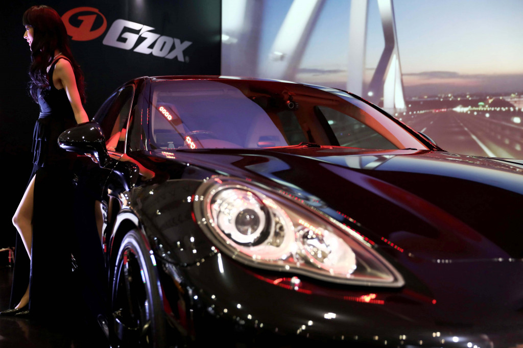 Seorang foto model berpose bersama Porsche yang tampil kemilau di sebuah booth produk perawatan mobil.