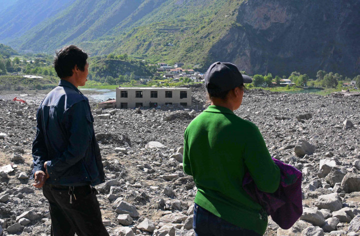 Warga menyaksikan sisa desa yang dilanda longsor. Bagian atas sebuah gedung nampak menyembul dari timbunan tanah berbatu yang menguburnya.