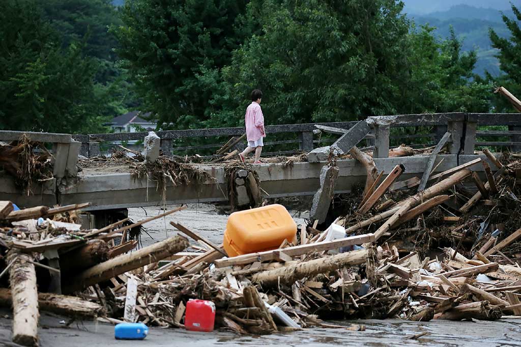 Seorang warga melewati jembatan yang rusak akibat banjir bandang, sementara puing-puing kayu tersangkut di bawah jembatan.
