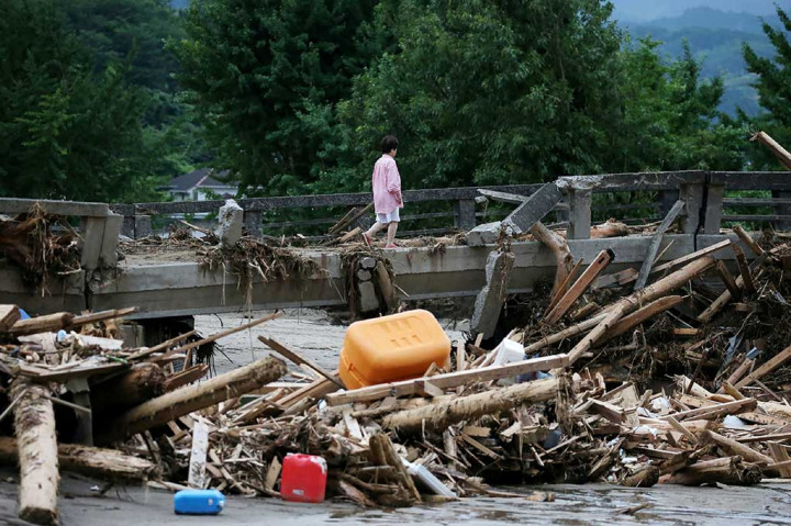 Seorang warga melewati jembatan yang rusak akibat banjir bandang, sementara puing-puing kayu tersangkut di bawah jembatan.