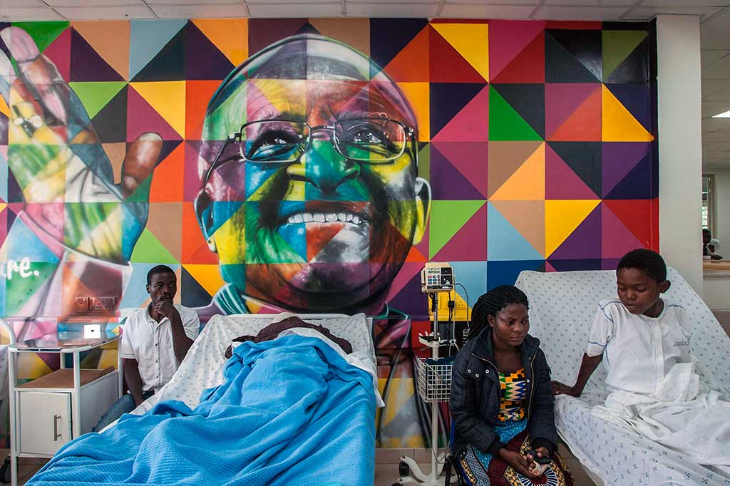 Pasien berbaring di tempat perawatan dengan latar belakang mural uskup Afrika Selatan Desmond Tutu.