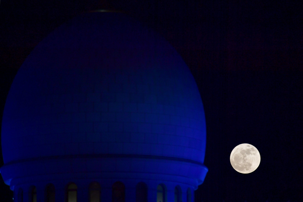 Fenomena supermoon terlihat di balik salah satu kubahnya. Cahaya biru dan putin disorotkan bergantian ke tembok masjid untuk menandai waktu salat.