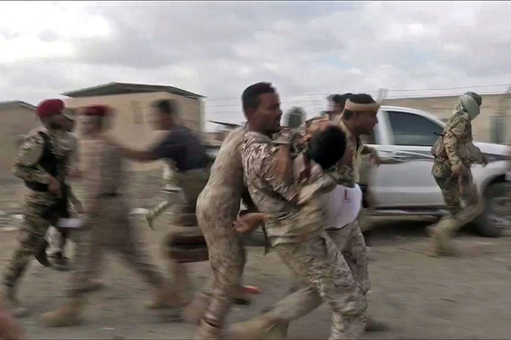 Tentara bergegas membawa kawan yang terluka ke kendaraan militer, sementara seorang pria memegang kamera berdarah di tanah.