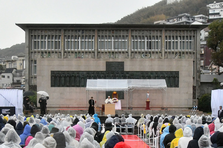 Ratusan orang rela diguyur hujan demi mendengarkan Fransiskus, yang berdiri di samping sebuah foto historis kengerian bom Nagasaki. 