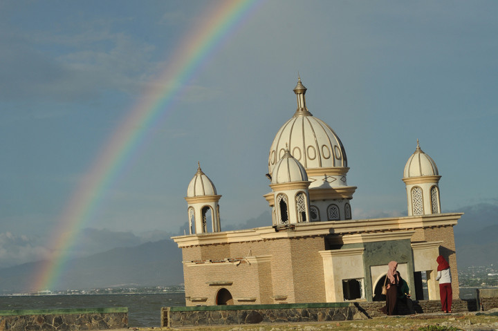 Sejumlah warga menikmati suasana menjelang matahari tenggelam dengan latar belakang pelangi serta masjid terapung yang rusak akibat gempa dan tsunami di Pantai Teluk Palu, Palu, Sulawesi Tengah, Selasa, 31 Desember 2019. Sebagian warga di Kota Palu merayakan pergantian tahun dengan menikmati suasana matahari terbenam terakhir tahun 2019 di pantai tersebut. Antara Foto/Mohamad Hamzah