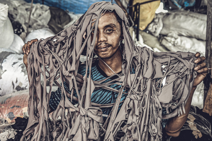 Seorang pengepul berpose dengan sisa kain tekstil yang telah terpotong-potong di tempat penampungan miliknya di kawasan Jakarta Utara. Potongan kain tersebut menjadi cerminan dari iklim perilaku kapitalis di industri 'fast fashion' dan budaya konsumtif manusia sehingga menyebabkan sampah over-konsumsi.