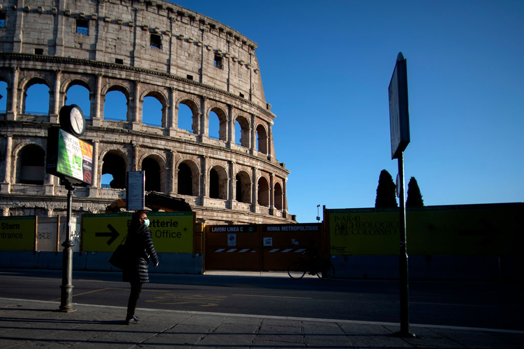 Seorang perempuan mengenakan penutup muka menunggu di halte bus di dekat monumen Colosseum, di sepanjang Via dei Fori Imperiali, Roma, Senin, 23 Maret 2020, selama lockdown yang bertujuan menghentikan penyebaran pandemi covid-19.