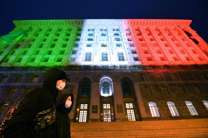 Warga yang memakai masker berjalan melewati Balai Kota Kiev, Ukraina yang diterangi warna bendera Italia, sebagai aksi solidaritas terhadap Italia yang saat ini paling terpukul oleh merebaknya covid-19. AFP PHOTO/Sergei Supinsky