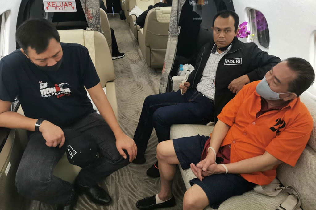 Di gambar lainnya Djoko Tjandra mengenakan baju tahanan dan tangan diborgol dikawal petugas kepolisian ketika berada di pesawat jenis Embraer Legacy 600 dengan nomor registrasi PK-RJP saat dijemput langsung oleh pihak berwajib di Malaysia.