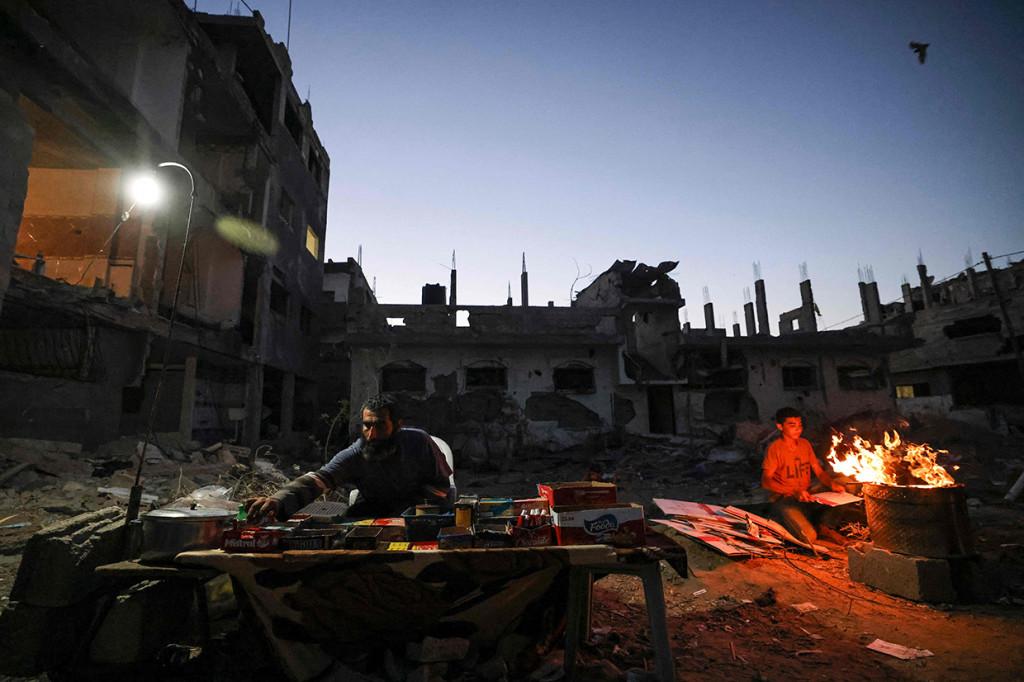 Seorang pria Palestina menjual barang-barang ketika seorang pemuda Palestina bersiap untuk memasak jagung dekat bangunan yang hancur selama konflik antara Hamas dan Israel pada Mei 2021 di Beit Hanun, Jalur Gaza, Senin, 7 Juni 2021.
