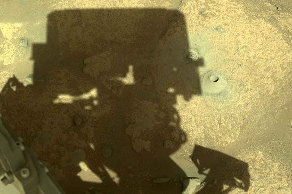 Bayangan penjelajah Mars Perseverance berada di sebelah gundukan kecil dengan lubang di tengahnya di planet Mars.