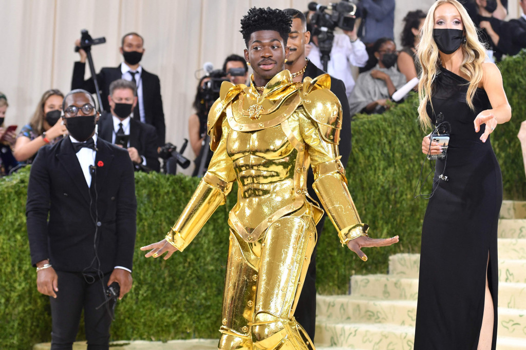 Lil Nas X mengenakan jubah yang menjuntai di karpet dari Atelier Versace berlapis emas. Di dalamnya, rapper ini mengenakan baju besi emas yang juga membalut gaya akhir, yakni catsuit berkilauan.
