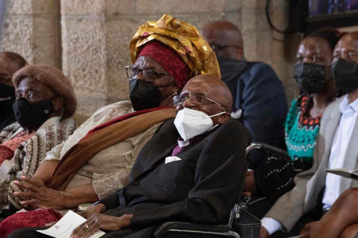 Tutu, mengenakan kemeja ungu khasnya dan kerah putih, di bawah setelan hitam, melambaikan tangan sebagai tanda terima, dari kursi roda.