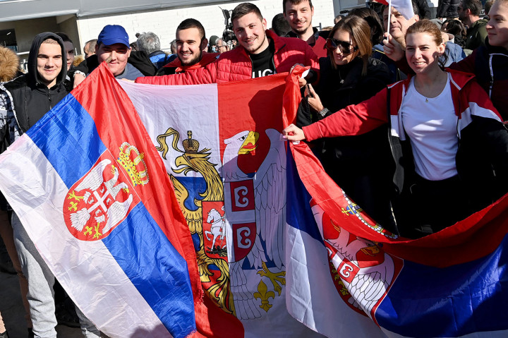 Orang-orang memegang bendera nasional Serbia saat mereka  menunggu di luar pintu keluar VIP bandara internasional Beograd pada 17 Januari 2022, untuk kedatangan petenis nomor satu dunia Novak Djokovic setelah dideportasi dari Australia karena status vaksinasi virus coronanya.