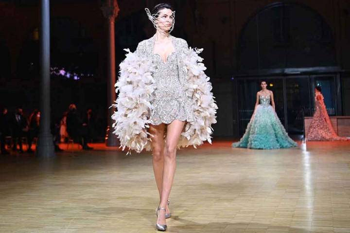 Model-model mengitari ruangan dengan gaun pesta tulle dan jubah yang dilapisi bunga bugenvil cerah, kontras dalam tekstur dan warna dengan gaun bernada biru mengilap yang dilapisi bordir.