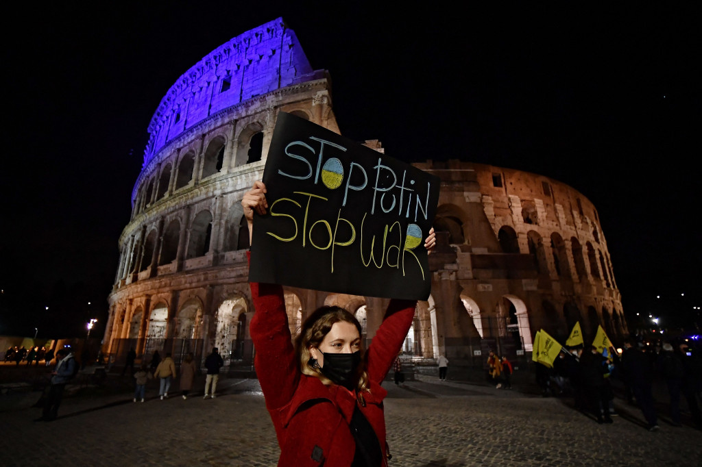Seorang pengunjuk rasa memegang spanduk di depan Colosseum Kuno, yang diterangi cahaya warna bendera Ukraina.