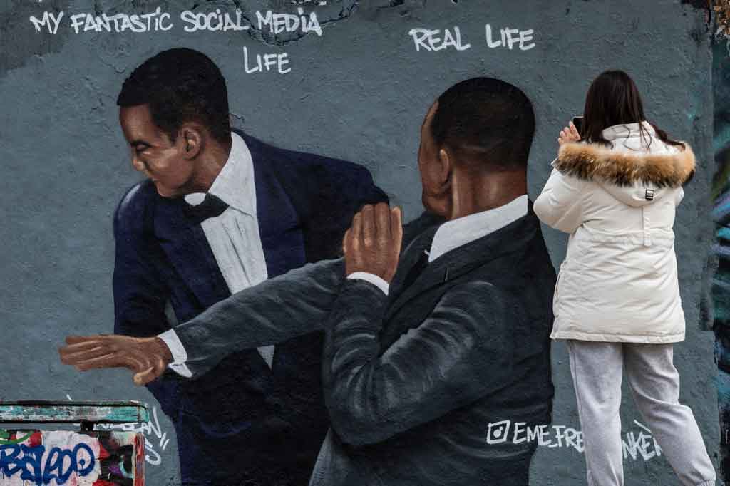 Seorang wanita mengambil foto lukisan dinding karya seniman jalanan yang berbasis di Berlin, Eme Freethinker, yang menampilkan aktor AS Will Smith (kanan) menampar komedian AS Chris Rock selama upacara Oscar, di Berlin pada 30 Maret 2022.