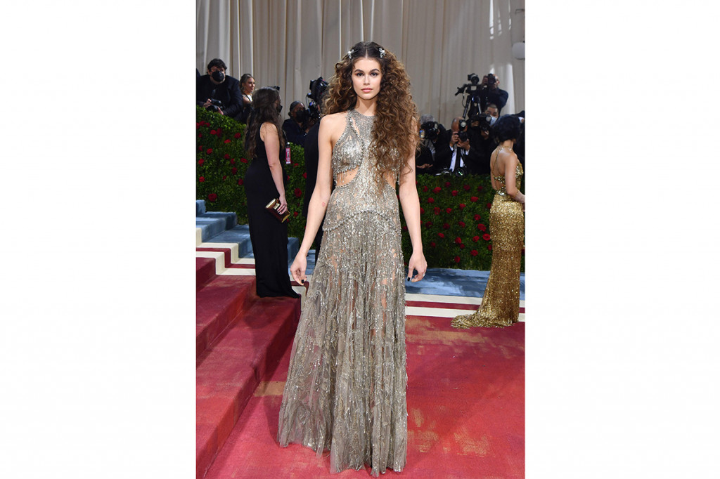 Model Kaia Gerber berpose dalam balutan gaun sheer silver dengan detail cut-out di kedua sisinya oleh Alexander McQueen. Rambutnya ditata dengan sentuhan curly dan menampilkan dua klip perak sebagai hiasannya.