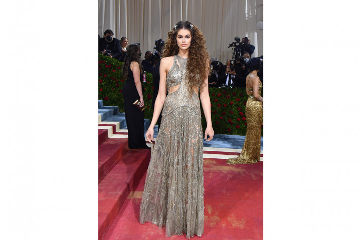 Model Kaia Gerber berpose dalam balutan gaun sheer silver dengan detail cut-out di kedua sisinya oleh Alexander McQueen. Rambutnya ditata dengan sentuhan curly dan menampilkan dua klip perak sebagai hiasannya.