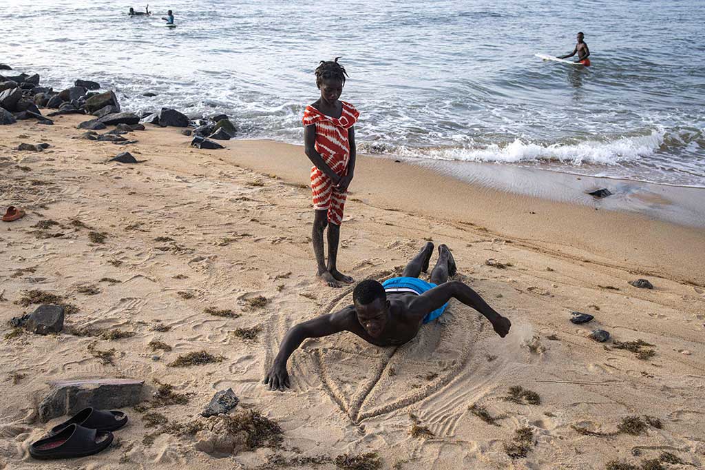 Di pantai yang dipenuhi bebatuan, Oscar, bertelanjang dada dan berbaring di pasir, menggunakan tangannya untuk menunjukkan kepada seorang gadis muda yang berdiri di sampingnya cara mendayung di air. Sapuannya yang lebar membuat bekas di pasir.