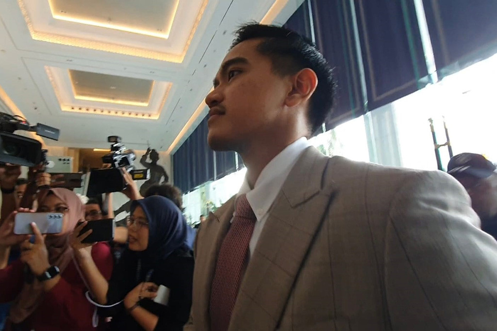 Kaesang memakai setelan jas cokelat. Lengkap dengan dasi merah muda.