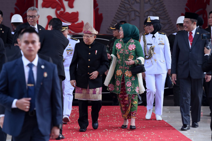 Wapres tampak mengenakan penutup kepala, kemudian beskap (jas), celana panjang, dan kain songket yang dipakaikan di pinggang hingga lutut.